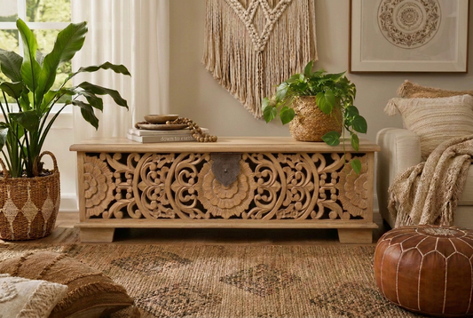 A carved wooden chest of drawers in a light weathered gray finish, styled in a cozy, bohemian-themed living room next to a sofa with a plant on top.